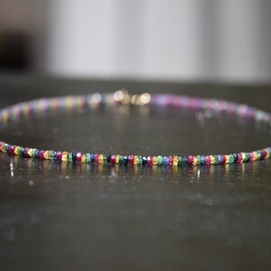 Pu&ograve; includere: Una delicata collana con una fila di piccole pietre preziose colorate. Le perline sono disposte in un motivo arcobaleno, con sfumature di rosso, giallo, verde, blu e viola. La collana ha una chiusura dorata.