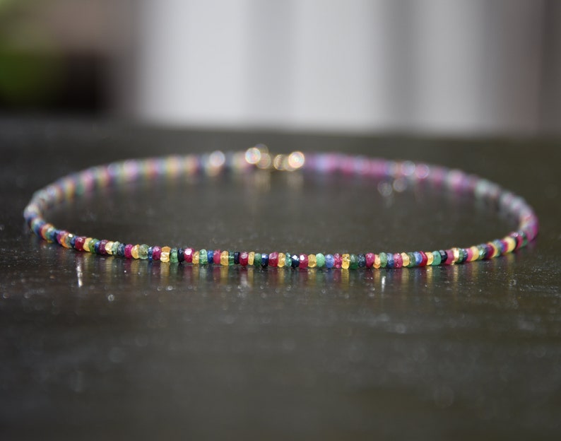 Pu&ograve; includere: Una delicata collana con una fila di piccole pietre preziose colorate. Le perline sono disposte in un motivo arcobaleno, con sfumature di rosso, giallo, verde, blu e viola. La collana ha una chiusura dorata.