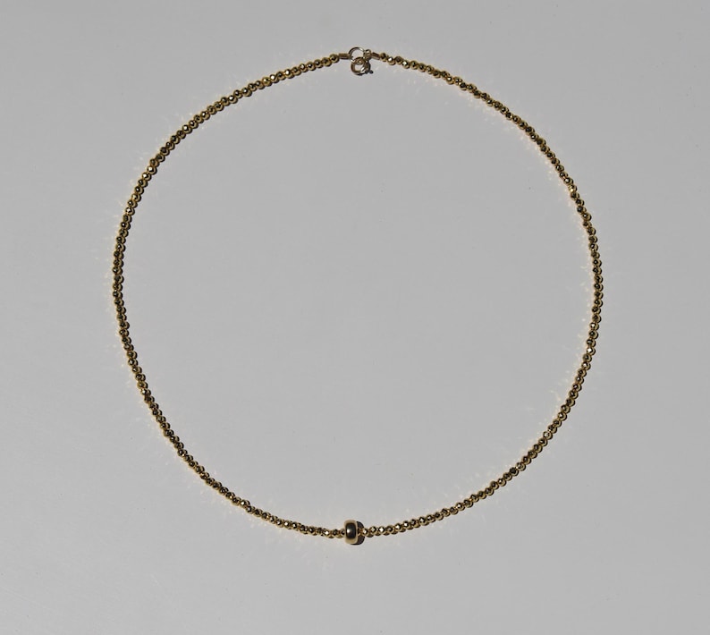 May include: A delicate gold necklace featuring a fine chain of small, round beads. The necklace has a simple design with a single, slightly larger bead near the center and a clasp closure. The jewelry is displayed against a plain white background.