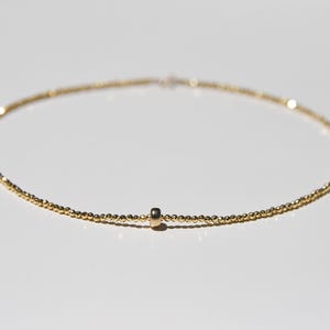 May include: Delicate gold necklace featuring a strand of small, round beads. A small, cylindrical gold charm is centered on the necklace. The necklace is displayed on a white surface, with soft lighting.