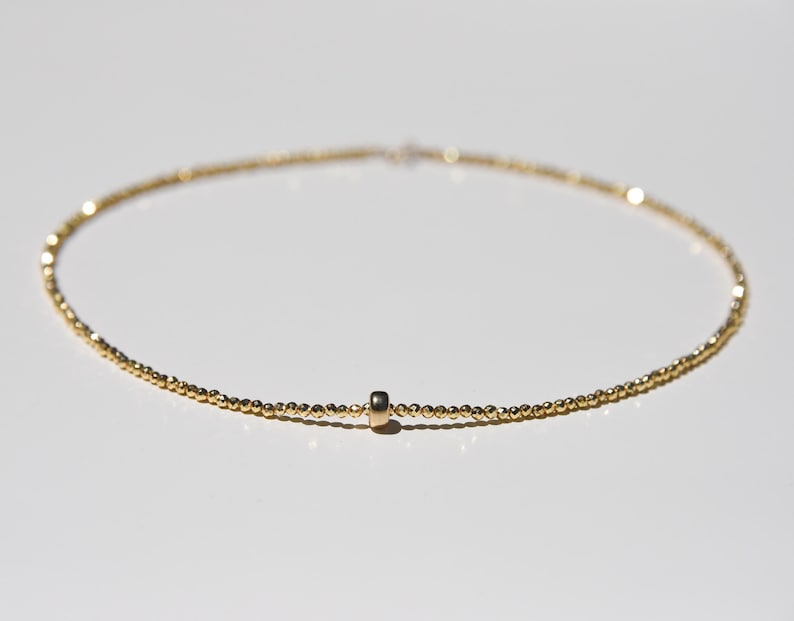 May include: Delicate gold necklace featuring a strand of small, round beads. A small, cylindrical gold charm is centered on the necklace. The necklace is displayed on a white surface, with soft lighting.