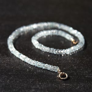 May include: A delicate aquamarine gemstone necklace. The necklace features small, faceted, light blue aquamarine beads strung together. The clasp is a small, gold-toned metal. The necklace is displayed on a dark surface.