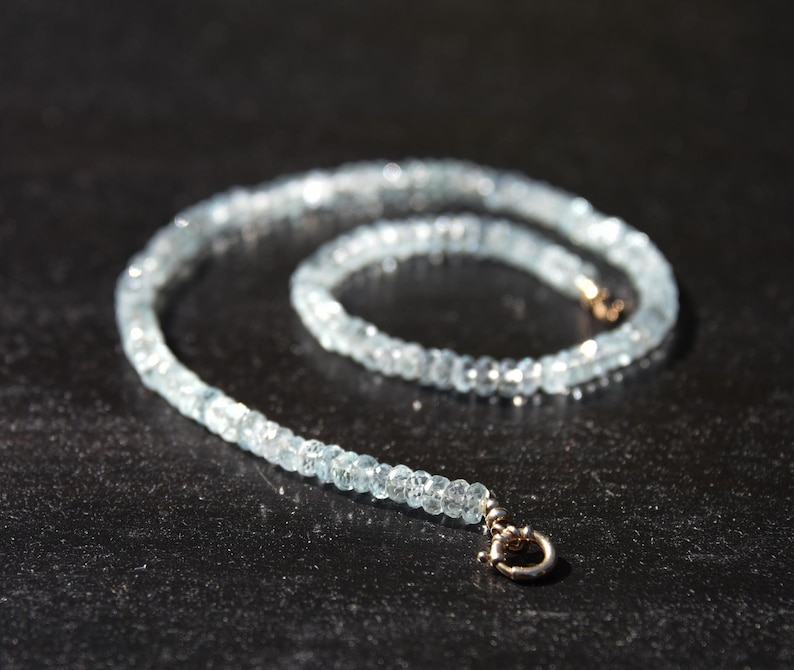 May include: A delicate aquamarine gemstone necklace. The necklace features small, faceted, light blue aquamarine beads strung together. The clasp is a small, gold-toned metal. The necklace is displayed on a dark surface.