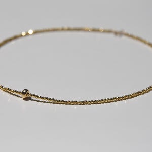 May include: Delicate gold necklace featuring a strand of small, evenly spaced beads. A single, larger bead is positioned near the clasp. The necklace is displayed on a white surface, highlighting its simple elegance.