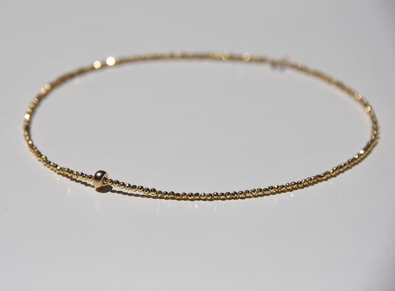 May include: Delicate gold necklace featuring a strand of small, evenly spaced beads. A single, larger bead is positioned near the clasp. The necklace is displayed on a white surface, highlighting its simple elegance.