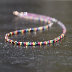 Pu&ograve; includere: Una collana colorata composta da piccole pietre preziose rotonde in un arcobaleno di colori. Le pietre sono infilate insieme, creando un accessorio delicato e vibrante. La collana &egrave; esposta su uno sfondo scuro.