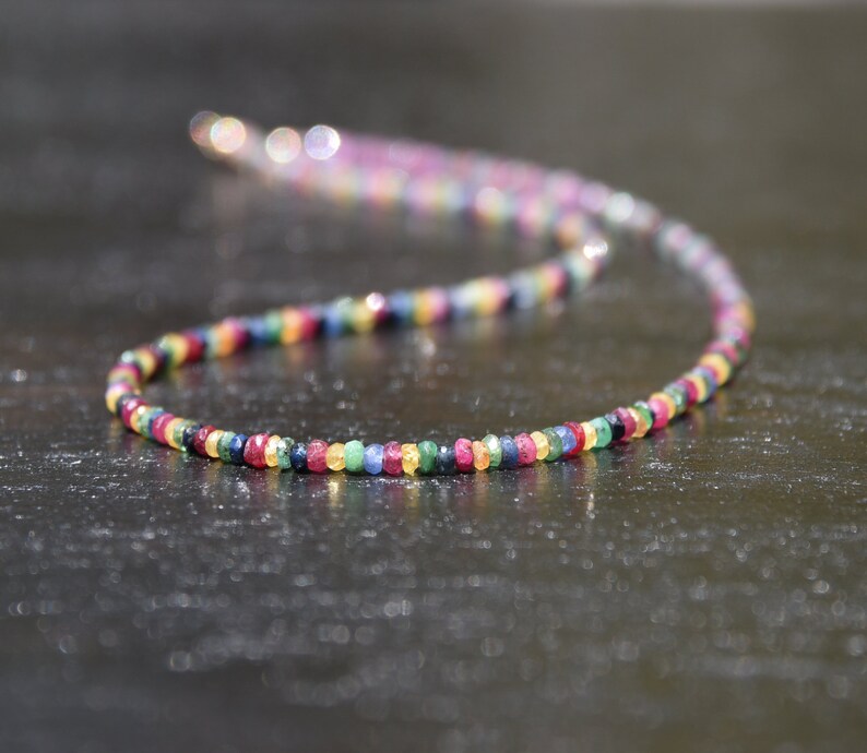 Pu&ograve; includere: Una collana colorata composta da piccole pietre preziose rotonde in un arcobaleno di colori. Le pietre sono infilate insieme, creando un accessorio delicato e vibrante. La collana &egrave; esposta su uno sfondo scuro.