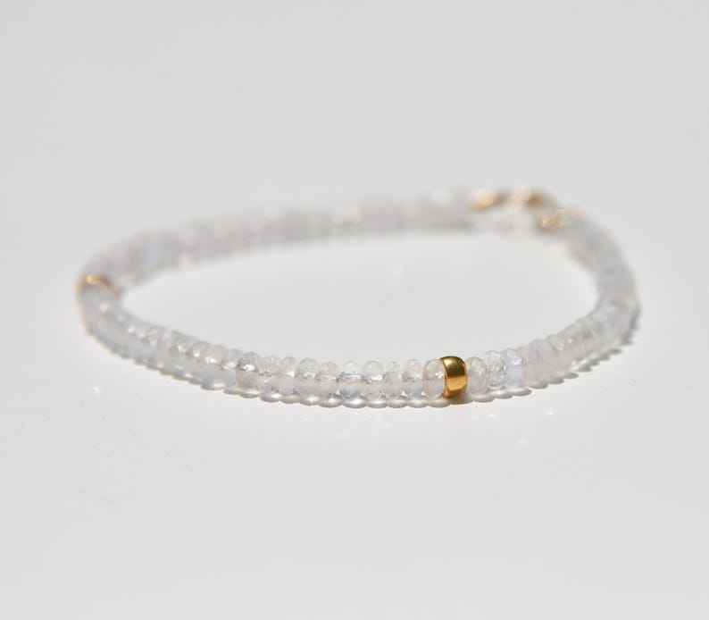 May include: Delicate white beaded bracelet with a gold accent bead. The bracelet is made of small, round, translucent beads strung together. The gold bead adds a touch of contrast and elegance to the minimalist design. The bracelet is shown on a white surface.
