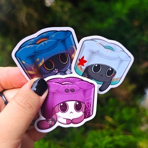 May include: Three cute cat stickers with different colored hats. The first sticker is blue with gold accents, the second is white with green accents, and the third is purple with black accents.