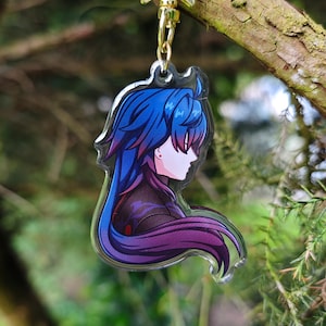 May include: A clear acrylic keychain featuring a purple-haired anime character with blue eyes. The character is depicted in profile, looking to the right. The keychain is hanging from a gold chain.