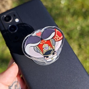 May include: A clear phone grip with a cartoon character design. The character is wearing a red and gold hat with a white plume, a purple mustache, and a white beard. The character is holding a red and gold object in their hand.