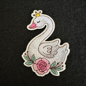 May include: A white embroidered swan with a gold crown and pink rose with green leaves. The swan is wearing a crown and has its eyes closed.