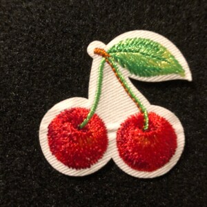 Op de afbeelding: Geborduurde kersenpatch. Twee rode kersen met groene stelen en een groen blad staan tegen een witte contour. De patch staat tegen een zwarte achtergrond. Ideaal om kleding of accessoires een fruitige touch te geven.