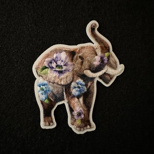 May include: Embroidered elephant patch with floral accents. The patch features a brown elephant adorned with purple and blue flowers. The patch is on a white background and is set against a black backdrop. This patch is ideal for adding a unique touch to clothing or accessories.