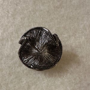 May include: A detailed, antique-style silver-toned button with a textured, radial design. The button has a three-dimensional, slightly curved shape with a decorative edge. The button's surface has a detailed, textured pattern.