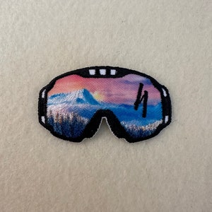 May include: Embroidered patch of ski goggles with a scenic mountain landscape reflected in the lenses. The patch features a black border and a colorful sunset scene with blue, pink, and yellow hues.