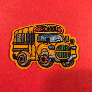 May include: Yellow school bus embroidered patch with the word "SCHOOL" on the front. The bus has black windows, a red light, and green grill.