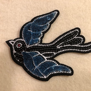 May include: A blue and black embroidered bird applique with a red eye. The bird is flying with its wings spread out.