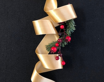 May include: A gold satin ribbon is curled and tied around a sprig of green pine with red berries.
