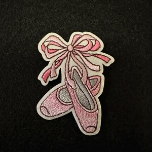 May include: Pink embroidered ballet shoe patch with a pink bow. The patch is on a white background.