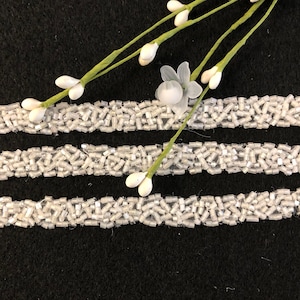 Pode incluir: Três fileiras paralelas de acabamento de contas brancas sobre um fundo preto. Delicados raminhos florais brancos com pequenos botões e flores são dispostos diagonalmente no topo. A guarnição parece ser feita de pequenas contas retangulares.