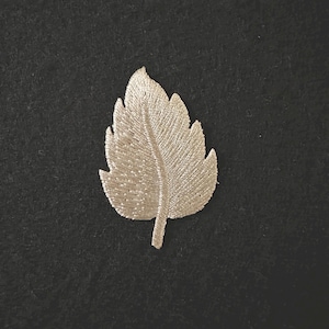 May include: A silver-colored embroidered leaf applique. The leaf has detailed stitching, with a central vein and side veins. The applique is set against a dark background, highlighting the intricate design. This item is suitable for sewing and craft projects.