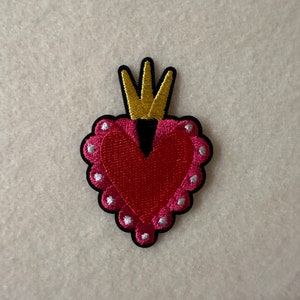 Puede incluir: Parche bordado con un corazón rojo coronado con una corona dorada. El corazón tiene un borde festoneado con puntos plateados y un borde negro. Para ropa o accesorios.