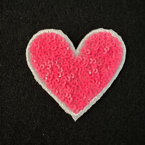 Könnte beinhalten: Ein pinkfarbener, paillettenbesetzter herzförmiger Aufnäher mit silbernem Rand. Das Herz ist mit kleinen, glänzenden Pailletten gefüllt. Der Aufnäher ist vor einem schwarzen Hintergrund platziert, was einen Kontrast erzeugt.