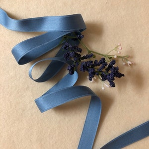 May include: A blue satin ribbon is curled on a beige background with a sprig of purple berries and small pink flowers.