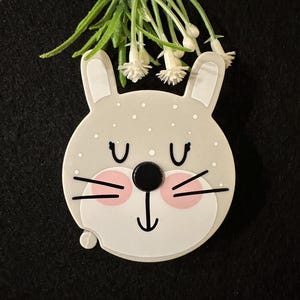 Puede incluir: Una cinta métrica en forma de conejo blanco y gris con ojos negros y mejillas rosadas. La cinta está decorada con lunares blancos. Una rama de flores blancas cuelga sobre la cinta.