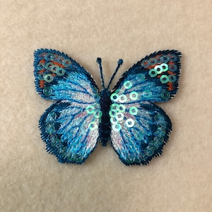 May include: A blue and white butterfly with iridescent sequins, embroidered on a fabric background.