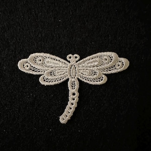 May include: A white lace dragonfly motif, featuring detailed wings and a slender body. The applique is set against a black background, accentuating the intricate design. Ideal for craft projects and embellishments. The dragonfly's wings are symmetrical.