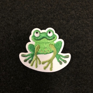 May include: Green embroidered frog patch with white stitching. The frog is sitting on a white background.