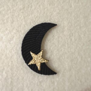 May include: A black crescent moon patch with a gold star attached. The moon is made of a textured, ribbed fabric. The star is a metallic gold color and is attached to the moon.