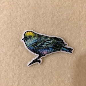 May include: A blue and green bird with a yellow head, embroidered on a white fabric patch.
