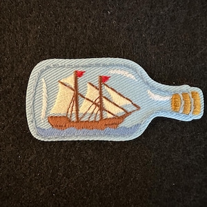 May include: Embroidered patch featuring a ship inside a bottle. The ship has brown masts and sails, with red flags. The bottle is light blue with a gold-colored neck, and the patch has a light blue border.