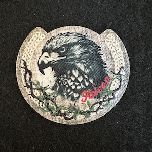 Pode incluir: Um patch circular com uma ilustração detalhada da cabeça de um falcão em tons de cinza e preto. A palavra "Falcon" é bordada em vermelho. O patch é cercado por uma coroa e uma borda texturizada.