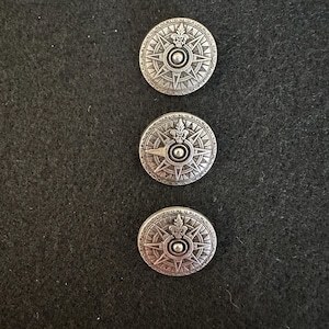May include: Three silver-coloured, round buttons with a compass rose design. Each button features a central circle, a fleur-de-lis, and a starburst pattern. The buttons are set against a dark background.