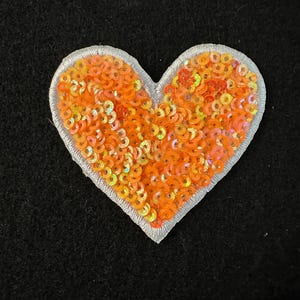 May include: A heart-shaped patch with orange sequins and a silver border. The sequins are densely packed, creating a shimmering effect. The patch is set against a black background, highlighting the vibrant colors.