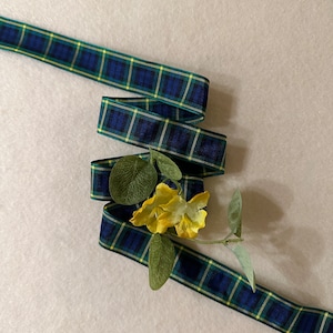 May include: A blue, green and yellow plaid ribbon is curled and tied in a bow. A small sprig of green leaves and yellow flowers is placed on top of the ribbon.