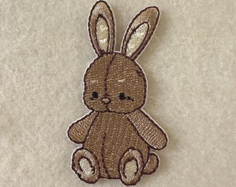 Może przedstawiać: Brązowa, haftowana naszywka z królikiem z białym obramowaniem. Królik ma długie uszy, małe czarne oczy i siedzi. Naszywka jest prawdopodobnie przeznaczona na odzież lub akcesoria.