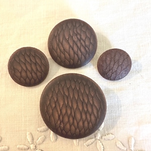 May include: Four circular buttons in varying sizes, each featuring a dark brown color and a raised, repeating scale-like texture across the surface. The buttons are arranged in a cluster, showcasing a consistent pattern of interlocking shapes.