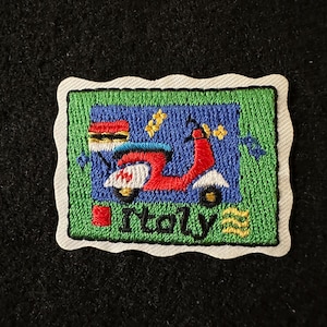 May include: Embroidered patch featuring a red scooter with white accents, set against a blue background. The patch is framed by a green border and the word "Italy" is displayed below the scooter. The patch has a white, wavy border.