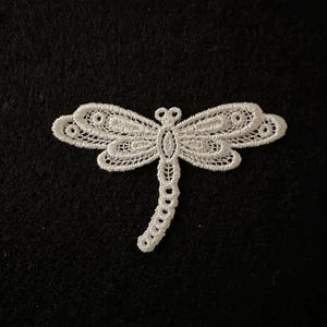 May include: A white, intricately detailed dragonfly applique on a black background. The dragonfly features delicate, openwork wings and a slender body, perfect for embellishing clothing or crafts. The design is symmetrical and elegant.