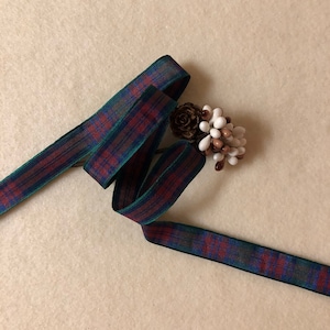 May include: A red, green, and blue plaid ribbon with a green edge. The ribbon is folded and twisted, and a small pine cone with white and brown beads is on top of the ribbon.