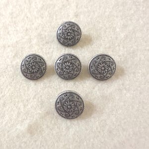 Puede incluir: Cinco botones redondos de color plateado con un diseño floral en relieve. Cada botón presenta una flor central rodeada de patrones arremolinados y un borde con cuentas. Los botones están dispuestos sobre una superficie de color claro.