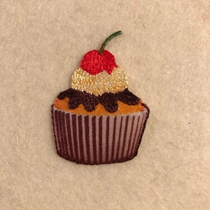 May include: A brown and white cupcake with chocolate frosting and a red cherry on top. The cupcake is embroidered on a fabric background.