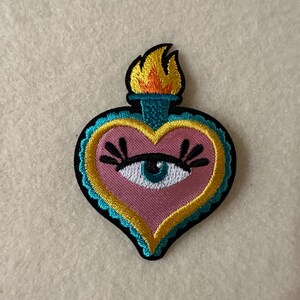 Puede incluir: Un parche bordado en forma de corazón rosa con un ojo azul y pestañas. El corazón está rodeado por un borde amarillo y azul y tiene una llama en la parte superior.