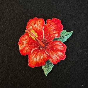 May include: Embroidered red hibiscus flower with white accents, yellow stamens, and green leaves. The floral design is set against a black background, showcasing the vibrant colours and intricate details of the embroidery.
