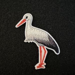 Op de afbeelding: Een witte en grijze geborduurde ooievaar met rode poten die op een zwarte achtergrond staat.